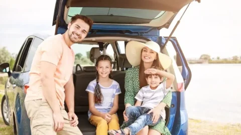 Güneşli bir günde, mavi bir arabanın arkasında poz veren mutlu bir aile. Anne ve baba, iki küçük çocuğuyla birlikte yaz tatilinin tadını çıkarıyor.